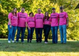A group of seven people, including Kim Stuebgen and her family, stand together outdoors wearing matching pink t-shirts that read "Every Pair Matters." They are smiling, surrounded by green trees and grass, at a breast cancer fundraiser held in Kim’s honor.