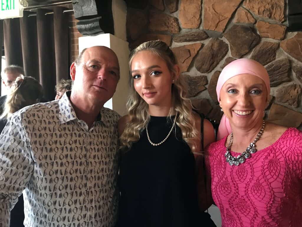 Three people pose together indoors: a man in a patterned shirt, a woman in a black dress with long blonde hair, and a woman in a pink headscarf and lace top. A stone wall is in the background.