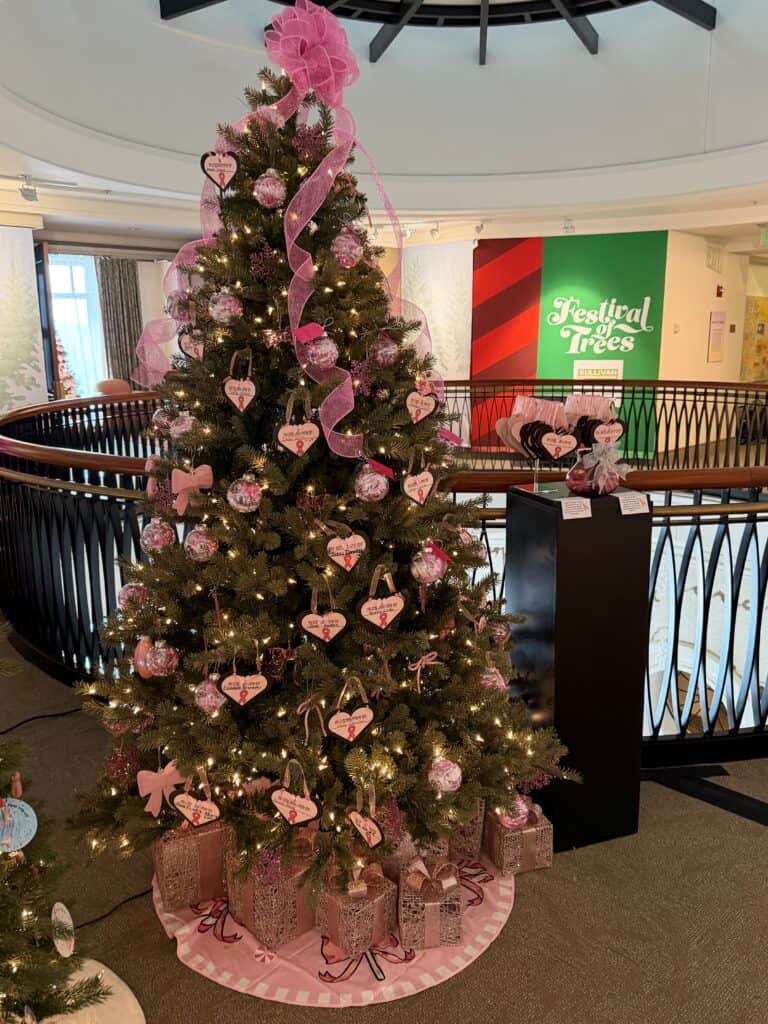 A Christmas tree decorated with pink ribbons, pink ornaments, and heart-shaped tags stands indoors, serving as a beautiful tree of hope. Pink-wrapped gifts are arranged underneath, while a green and red “Festival of Trees” sign in the background highlights this meaningful breast cancer fundraiser.