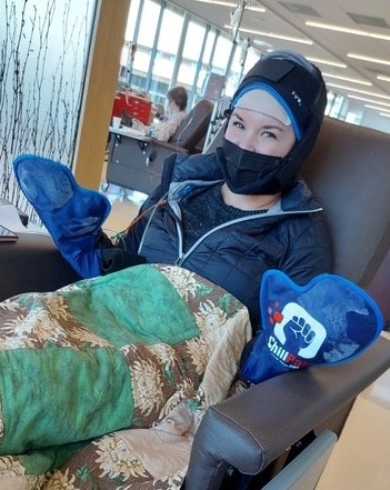 A woman sits in a recliner wearing a cold cap and mittens, covered with a green and brown floral quilt, in what appears to be a medical clinic. She is smiling and wearing a partial face mask.