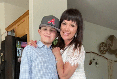 A single mom with dark hair in a white lace top and jeans smiles, hugging a boy in a checked shirt and baseball cap. They stand together in a cozy kitchen with wall decorations, showing strength through her metastatic breast cancer journey.