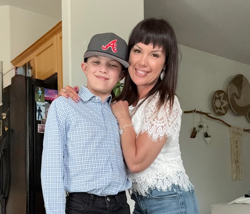 A single mom with dark hair in a white lace top and jeans smiles, hugging a boy in a checked shirt and baseball cap. They stand together in a cozy kitchen with wall decorations, showing strength through her metastatic breast cancer journey.