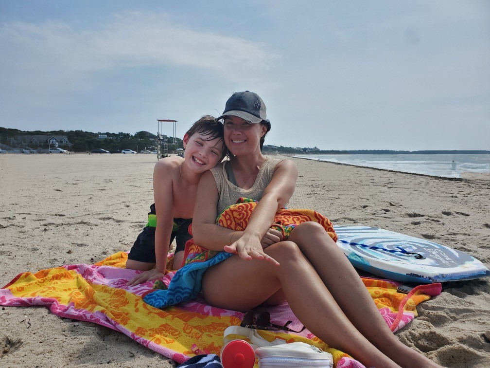 A smiling woman and a child sit on colorful towels at a sandy beach. The woman wears a hat and sunglasses, while the child hugs her from behind. A boogie board and beach items are nearby, with the ocean and sky in the background.