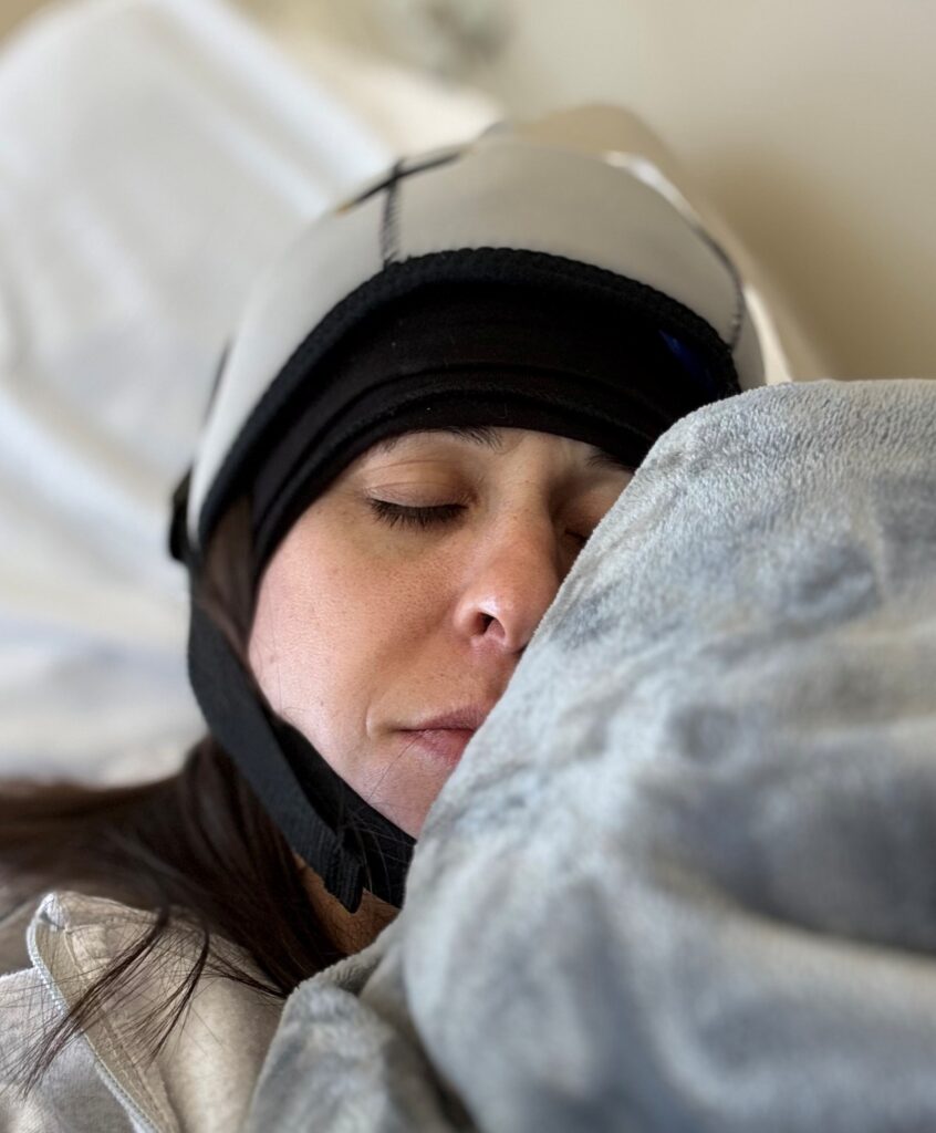 A young woman with breast cancer, wearing a cooling cap, rests with her eyes closed, lying in a hospital bed and wrapped in a gray blanket.