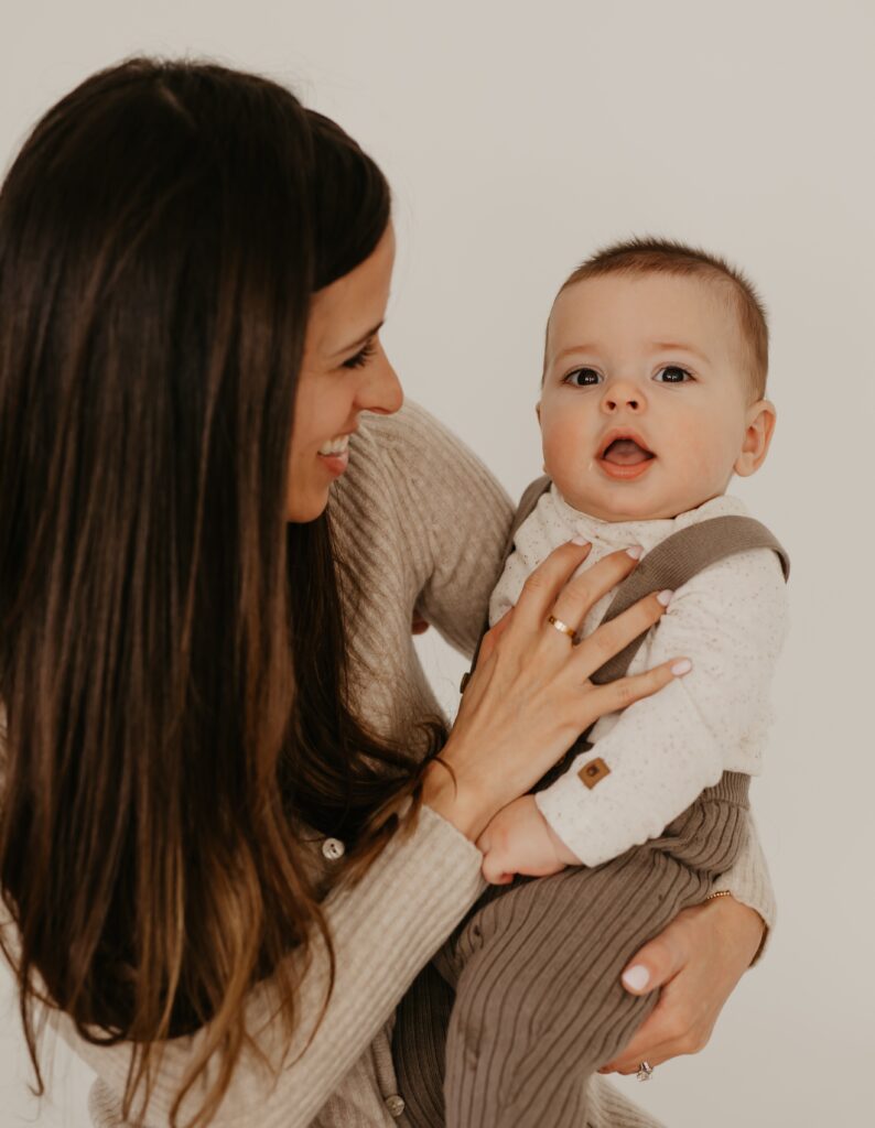 A woman with long brown hair smiles while holding a baby dressed in a light shirt and brown overalls. The baby looks at the camera with an open mouth, and the background is plain and light-colored.