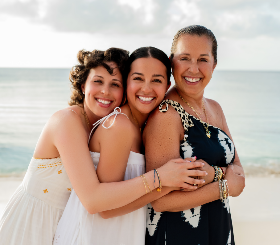 Three women smiling and hugging on a beach, wearing summer dresses and jewelry, with the ocean and cloudy sky in the background.