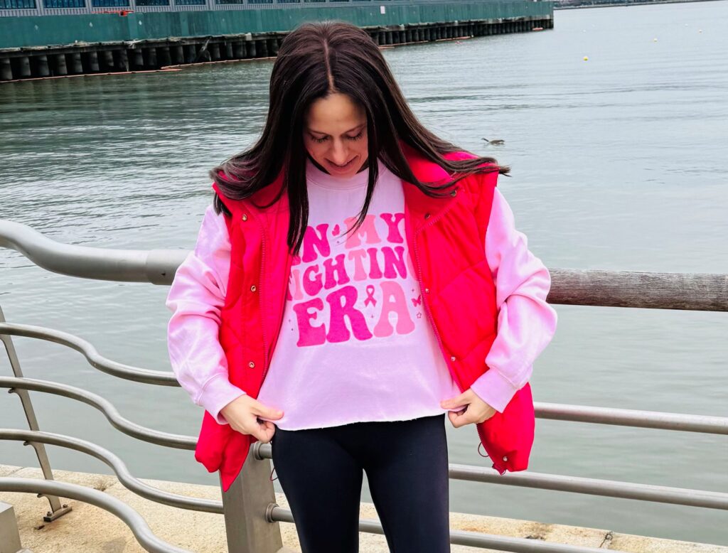 A young woman with breast cancer stands in front of a metal fence by a body of water. She is looking down and holding her pink shirt that reads "IN MY FIGHTING ERA."