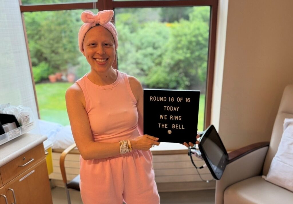 A smiling young woman with breast cancer in a pink outfit and headband holds a sign reading “ROUND 16 OF 16 TODAY WE RING THE BELL” in a bright room with large windows and greenery outside.