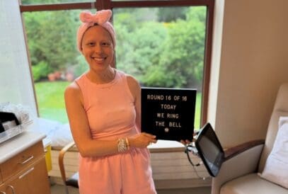 A smiling young woman with breast cancer in a pink outfit and headband holds a sign reading “ROUND 16 OF 16 TODAY WE RING THE BELL” in a bright room with large windows and greenery outside.