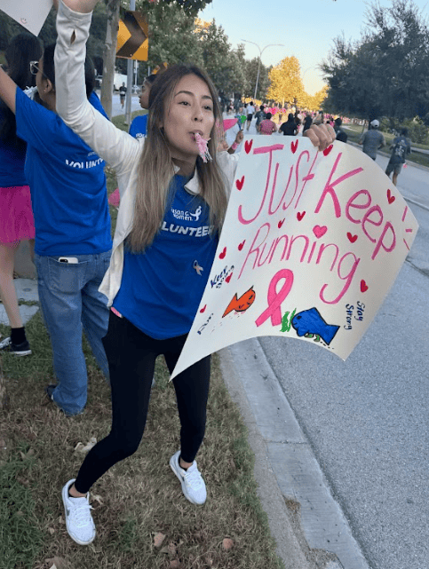 A woman in a blue volunteer shirt cheers and blows a whistle while holding a colorful sign that says “Just Keep Running” at an breast cancer fundraiser 5K event. Other participants are visible in the background.