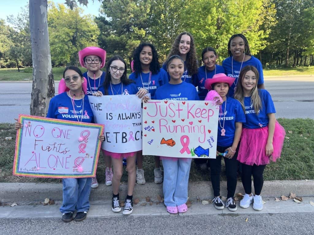 A group of young women in blue "Volunteer" shirts stand outdoors holding handmade signs supporting breast cancer awareness, including messages like "No One Fights Alone" and "Just Keep Running." Some wear pink accessories.