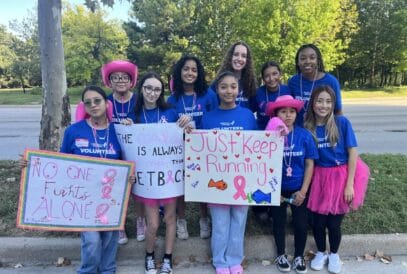 A group of young women in blue "Volunteer" shirts stand outdoors holding handmade signs supporting breast cancer awareness, including messages like "No One Fights Alone" and "Just Keep Running." Some wear pink accessories.