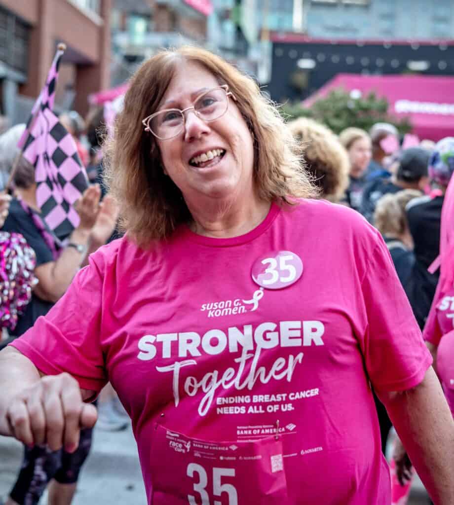 A smiling woman wearing glasses and a pink "Stronger Together" Susan G. Komen shirt stands among a group at a Race for the Cure event. She wears a pin with the number 35, which represents her 35 years as a breast cancer survivor.
