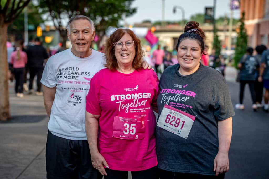 Three people stand together outdoors at a Race for the Cure event, all smiling. They wear race shirts and bibs; the two women’s shirts read “Stronger Together.” Other participants and trees are visible in the background.
