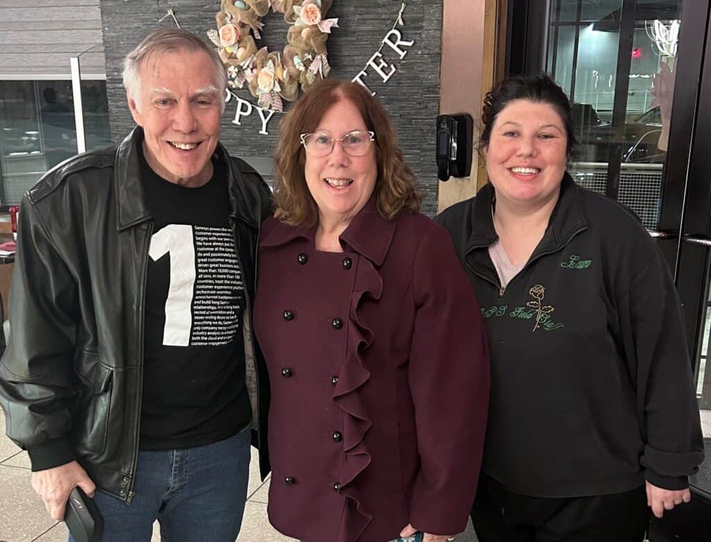 Three people pose and smile indoors. The man on the left wears a black jacket, the woman in the middle wears glasses and a maroon coat, and the woman on the right wears a black zip-up. A floral wreath and "Happy Easter" are in the background.