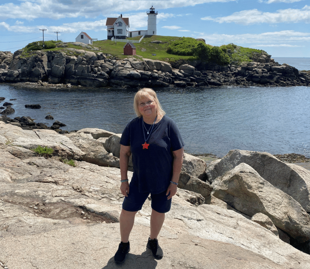 A blond woman in a blue shirt and shorts stands on rocky terrain by the water with a lighthouse and a small house visible on a hill in the background under a blue sky.