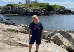 A blond woman in a blue shirt and shorts stands on rocky terrain by the water with a lighthouse and a small house visible on a hill in the background under a blue sky.