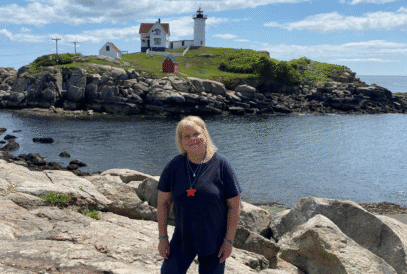 A blond woman in a blue shirt and shorts stands on rocky terrain by the water with a lighthouse and a small house visible on a hill in the background under a blue sky.
