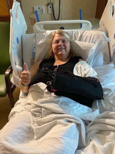 A woman with blond hair and glasses smiles from a hospital bed, giving a thumbs up to indicate she is feeling all right. She wears a black arm sling, indicating a broken arm.