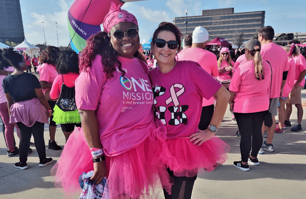 Two women in pink outfits, including T-shirts and tutus, pose at a Komen MORE THAN PINK breast cancer awareness walk. they smile broadly, surrounded by other participants in a sunny outdoor setting.
