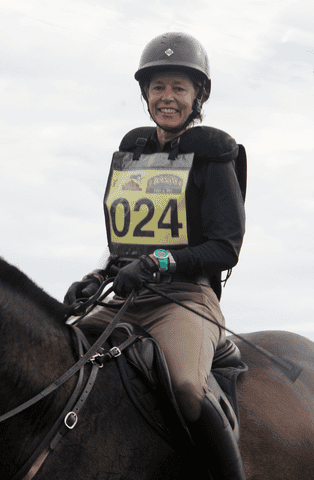 Nancy sits on a horse. She is wearing a helmet and holding a riding crop. She is participating in a competition.