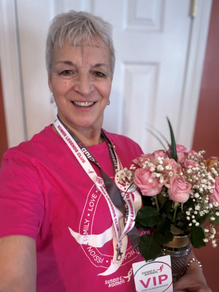 Smiling older woman with short gray hair wears a pink Susan G. Komen shirt and several lanyards, holding a bouquet of pink roses and a VIP Fundraising Legends Club badge. She stands in front of a white door.