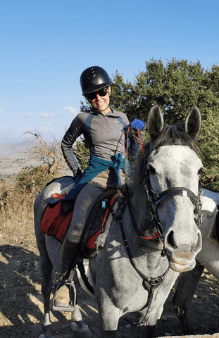 Breast cancer survivor Nancy is riding a large white horse. There is blue sky behind her. She is smiling