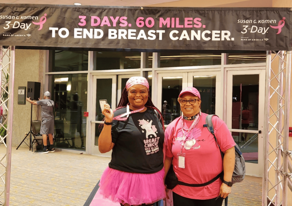 Two women in pink outfits smiling at a Susan G. Komen 3-Day walk to end breast cancer, standing under a banner reading "3 days. 60 miles. to end breast cancer."