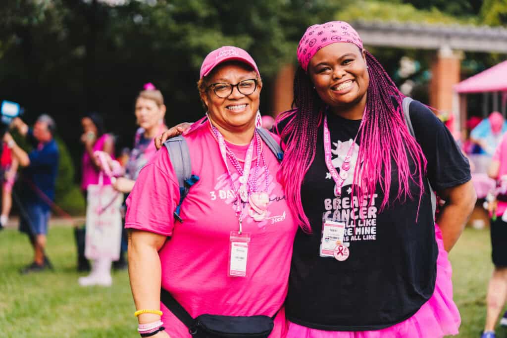 Two women dressed in pink, with matching accessories, smiling at a Komen 3-Day breast cancer awareness event. One wears glasses and a cap, and the other a bandana. they both have event badges.
