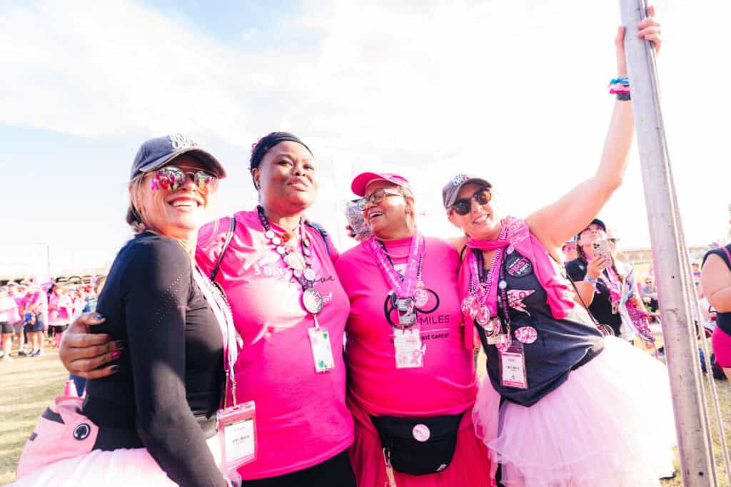 Four joyful women in pink attire, including tutus and sashes, celebrate at an outdoor event, embracing and smiling near a pole under a clear sky.
