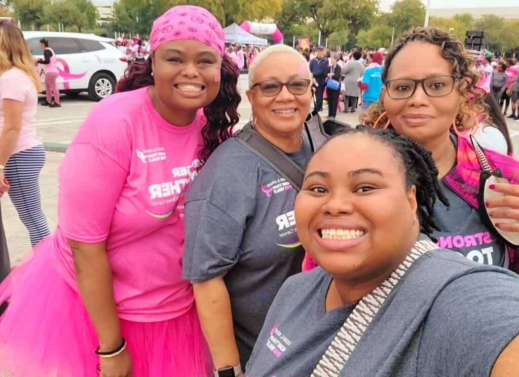 Four smiling women wearing pink outfits take a selfie at a Komen MORE THAN PINK Walk. One woman is in a pink tutu, and the background shows other participants and a pink-themed vehicle.