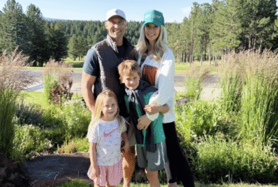 A smiling family of four, two adults and two children, pose together outdoors in front of tall grass and trees on a sunny day. The adults wear casual clothes and hats; the kids are dressed in sporty outfits.