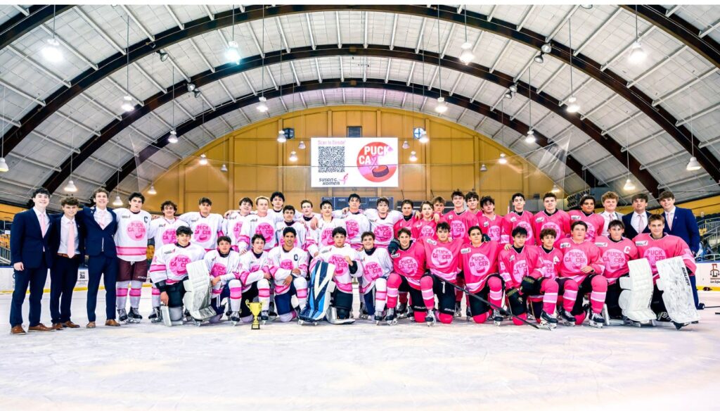 A large group of hockey players and coaches pose on an ice rink, wearing pink and white jerseys with “PUCK CANCER” logos, under an arched roof with bright lights, proudly supporting a hockey breast cancer fundraiser.