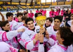A joyful hockey team in pink and white uniforms celebrates on the ice, holding a gold trophy together after their successful hockey breast cancer fundraiser. Teammates smile and reach for the cup inside an indoor rink with yellow seats in the background.
