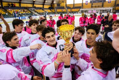 A joyful hockey team in pink and white uniforms celebrates on the ice, holding a gold trophy together after their successful hockey breast cancer fundraiser. Teammates smile and reach for the cup inside an indoor rink with yellow seats in the background.
