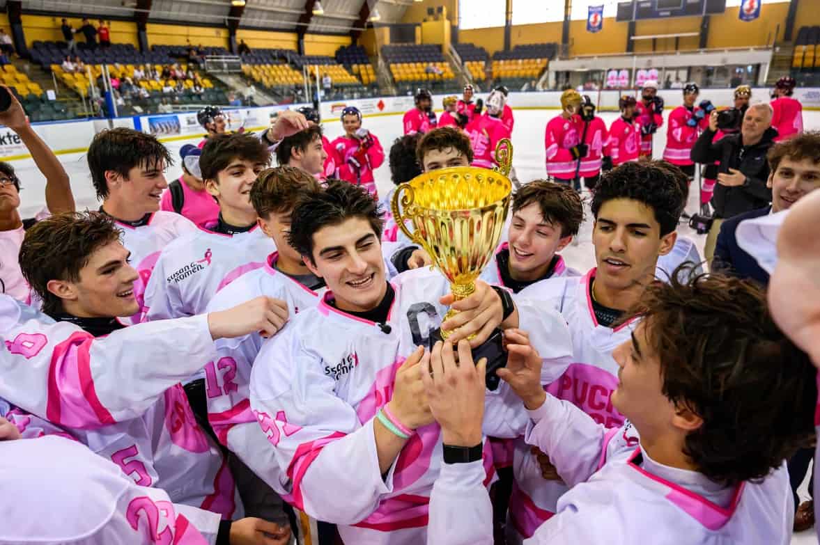 A joyful hockey team in pink and white uniforms celebrates on the ice, holding a gold trophy together after their successful hockey breast cancer fundraiser. Teammates smile and reach for the cup inside an indoor rink with yellow seats in the background.