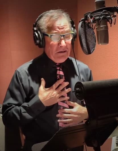A man with glasses is in a recording studio, wearing headphones. Donning a pink tie, he stands before a microphone, his hand to his chest as he reads from a script on a music stand. The backdrop of soundproofing panels attests to the importance of his message for the breast cancer ambassador fundraising campaign.