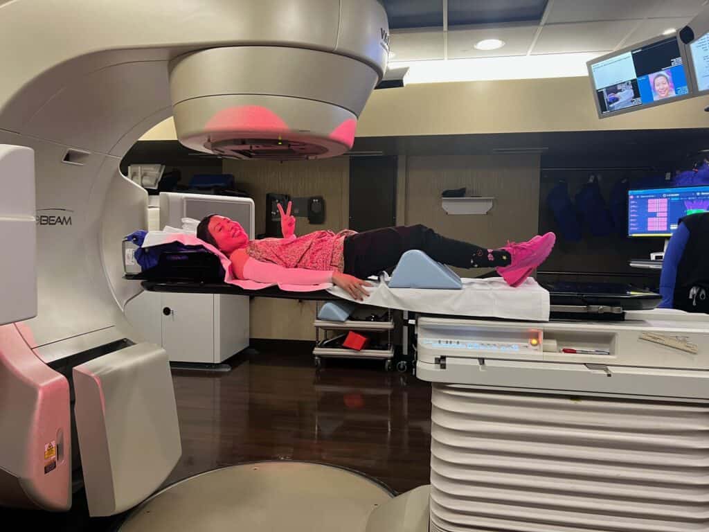 A person lies on a treatment table under a large medical machine for radiation therapy to treat breast cancer. The person wears pink clothes and shoes and flashes a peace sign, while medical equipment surrounds them.