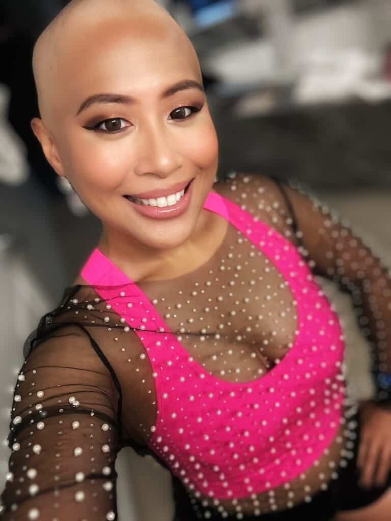 A smiling person with a bald head wearing a sheer black top with pearl embellishments over a bright pink tank top, taking a selfie indoors.