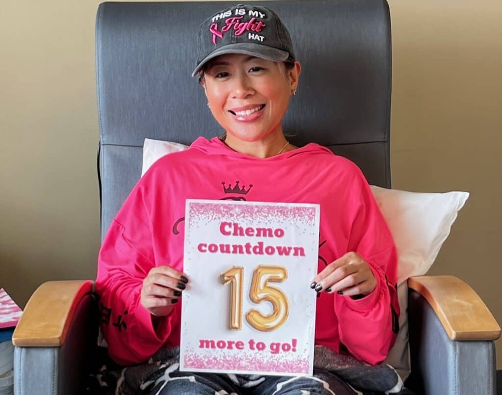 A smiling young woman with breast cancer wearing a pink shirt and a cap sits in a chair, holding a sign that reads, "Chemo countdown: 15 more to go!" The sign has pink borders and large gold numbers.