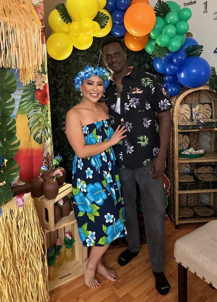 A smiling couple in Hawaiian-themed outfits stands together at a tropical-themed party with colorful balloons, palm leaves, and festive decorations in the background.