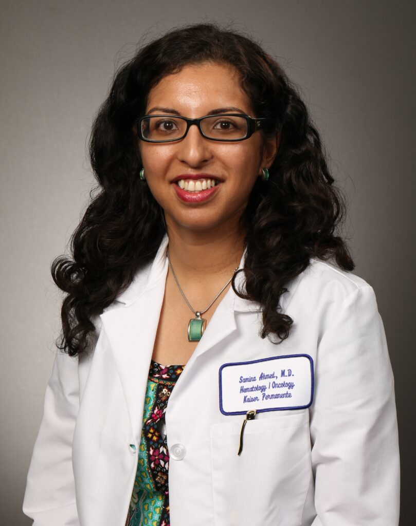A woman with curly dark hair and glasses smiles, wearing a white lab coat with a name tag that reads “Samina Ahmed, M.D., Hematology/Oncology, Kaiser Permanente,” and a turquoise necklace.