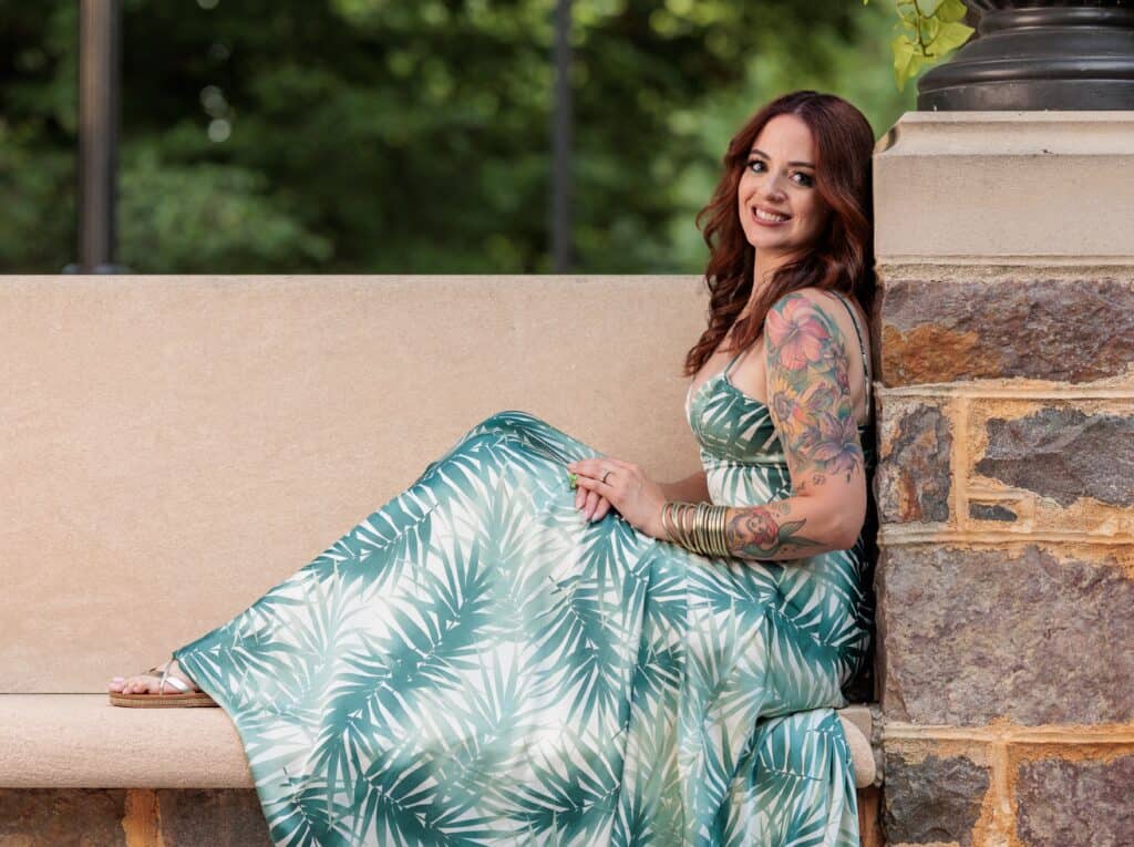 A woman in a green and white leaf-patterned dress sits on a stone bench, smiling, with her tattooed arm visible. Green trees are blurred in the background.