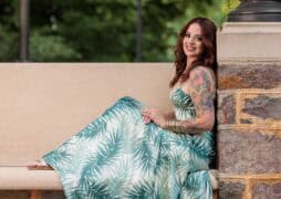 A woman in a green and white leaf-patterned dress sits on a stone bench, smiling, with her tattooed arm visible. Green trees are blurred in the background.
