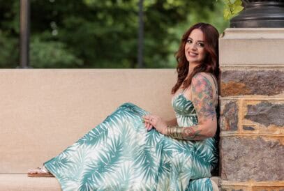 A woman in a green and white leaf-patterned dress sits on a stone bench, smiling, with her tattooed arm visible. Green trees are blurred in the background.
