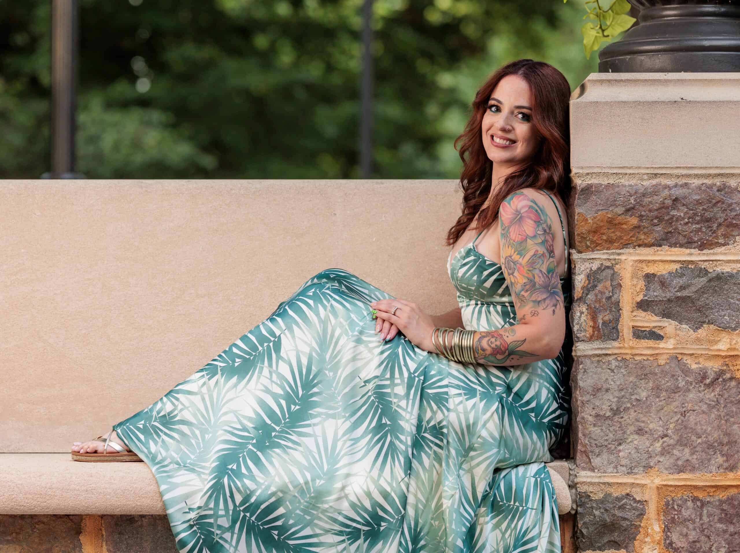 A woman in a green and white leaf-patterned dress sits on a stone bench, smiling, with her tattooed arm visible. Green trees are blurred in the background.