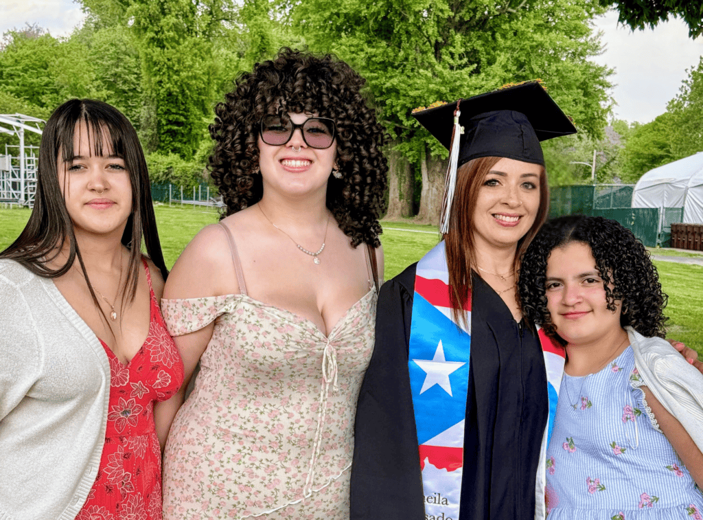Four women stand together outdoors, one in a graduation cap and gown with a Puerto Rican flag stole. They are smiling, with trees and grass in the background.