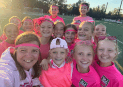 A group of smiling girls in pink athletic clothes and matching headbands pose closely together for a selfie on a sports field during daylight, celebrating their soccer breast cancer fundraiser.