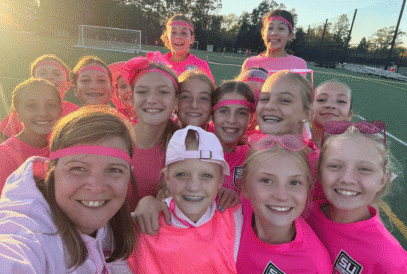 A group of smiling girls in pink athletic clothes and matching headbands pose closely together for a selfie on a sports field during daylight, celebrating their soccer breast cancer fundraiser.