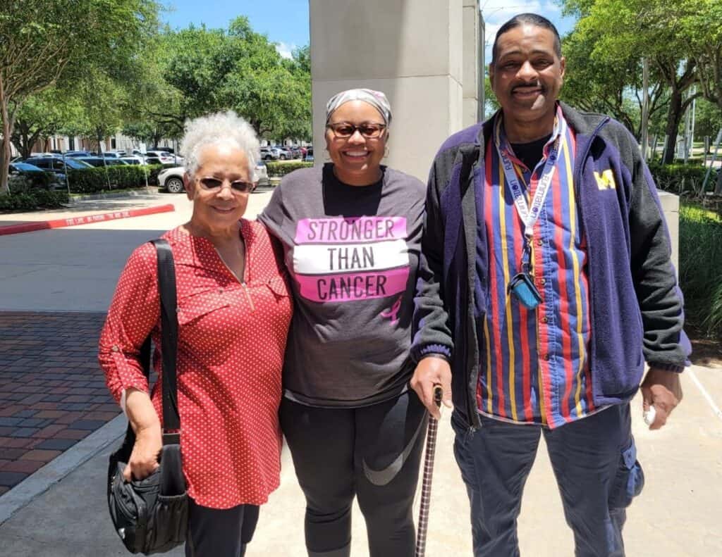 Three adults stand smiling outdoors on a sunny day. The person in the center, living with metastatic breast cancer, wears a headscarf and a "Stronger Than Cancer" shirt as their companions stand close, showing support. Trees and parked cars are behind them.
